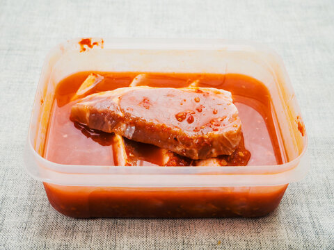 Plastic container with marinated pork chops on a light color table cloth surface. Meat in spicy red oriental souse. Food preparation.
