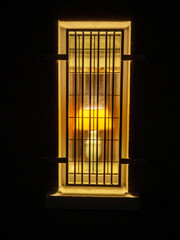 A window with a lamp in it is lit up in the dark. The light from the lamp shines through the window, creating a warm and cozy atmosphere. There is steel security protection.