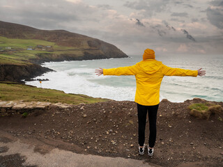 Woman tourist in yellow hat and jacket looking at stunning rough stone coastline in county Kerry,...