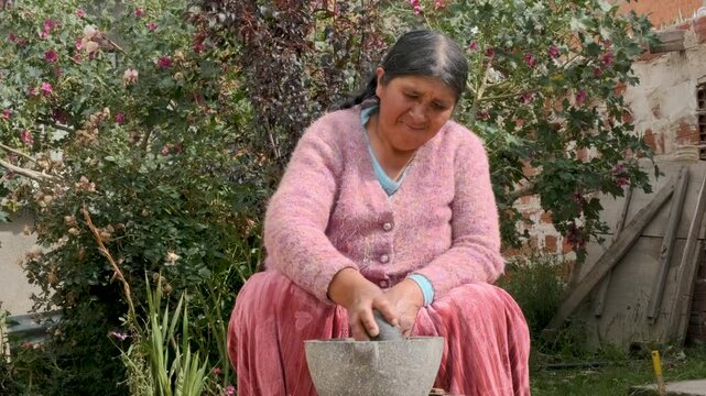 old woman of the aymara chola culture grinding in a mortar or also called batan in the courtyard of her house in la paz, bolivia - cooking concept