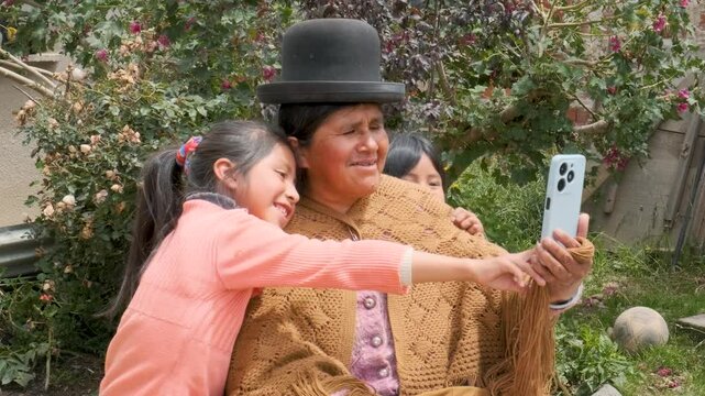 aymara cholita grandmother in pollera taking a selfie with her cell phone with her two granddaughters in the courtyard of her house in the city of la paz bolivia - communication concept