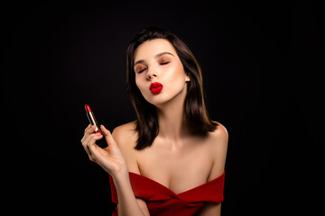 Obraz premium Gorgeous young woman in red dress with makeup holding lipstick against dark background