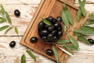 Board with bowl of tasty black olives and leaves on light wooden background