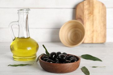 Bowl with tasty black olives and jug of oil on white wooden background