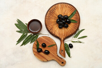 Wooden boards with tasty black olives, leaves and bowl of oil on light background