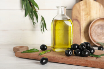 Board with tasty black olives and jug of oil on white wooden background