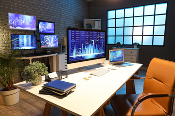 Computer and laptop with stock data on trader's desk in dark office at night