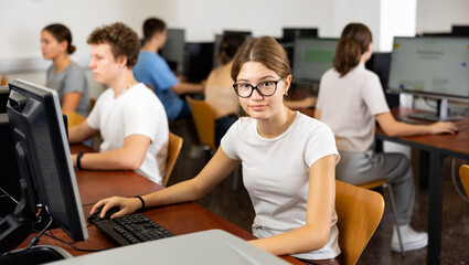 Portrait of positive smart girl in glasses sitting at desk in computer class, smiling and looking at camera.