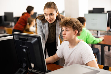 Female teacher and her student, teenage boy, looking at monitor of PC during computer science lesson.