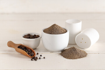Bowl of black pepper powder and scoop with peppercorns on white wooden background