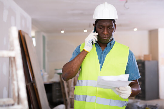 African american foreman with documents in his hands is talking on a mobile phone, discussing the workflow on the construction ..site indoors