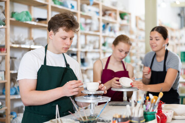 Young woman teacher in apron teaches teenage boy and girl students to make ceramic cup in ceramic workshop