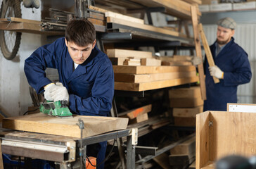 Young male carpenter polishes wooden plank with polishing machine in wood workshop