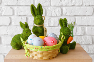 Trhee decorative green moss bunny figures with colorful Easter eggs in wicker basket on wooden table on white brick wall background. Easter holiday festive atmosphere, wallpaper, backdrop.