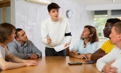Focused young guy student holding papers ad discussing assignments or projects with interested male group of various ages and nationalities gathered around table in classroom..