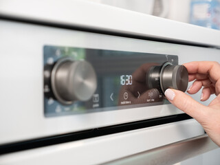Woman adjusting modern white oven in kitchen.  Female hand pushing and turning knobs on oven control panel