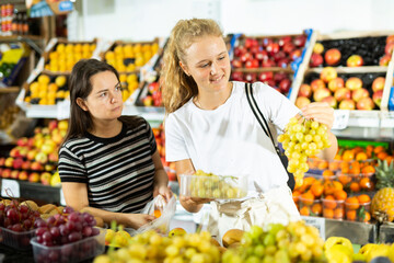 Young woman with a fifteen-year-old girl, who came to the store, attentive choose grapes, standing near the fruit counter