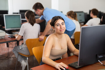 Fototapeta premium Positive teenage girl sitting at table and using computer during lesson.
