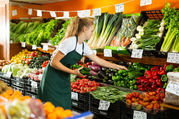 Positive young female seller in uniform holding green pepper in grocery shop