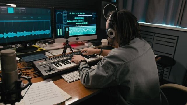 Medium rear shot of young Caucasian male composer sitting in recording studio in front of computer screens, playing chords on synthesizer while working on new song, sheet music on desk