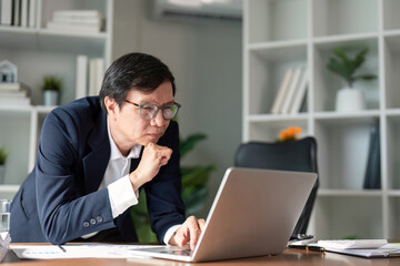 Elderly businessman deep in thought while working on a laptop in a modern office.