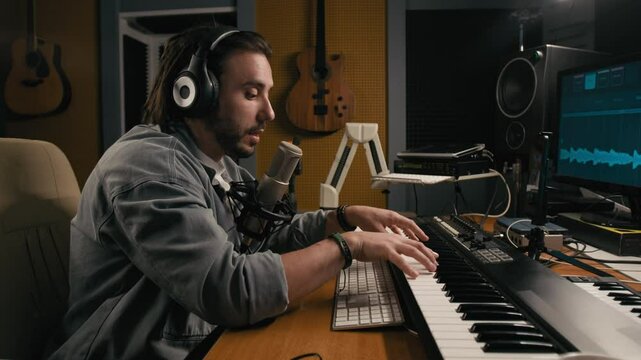 Medium side shot of cheerful young Caucasian male composer in headphones playing music on electronic synthesizer and speaking into microphone, while conducting masterclass or recording podcast