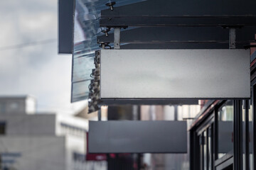 An empty white rectangular sign hangs outside a modern storefront in Germany. The metal frame is seen from below, leaving space for potential branding or advertisement in an urban environment.
