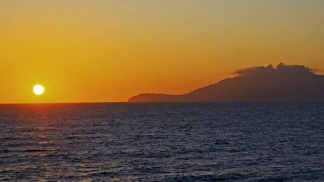 Setting sun just above ocean horizon paint the sky rich colors of orange, amber and gold-Distant island points the eye toward the beautiful sunset