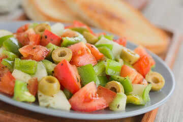 Fresh vegan salad of tomato, olive, green pepper and cucumber, seasoned with oregano, salt, black pepper and olive oil, served on blue plate with toasted bread in the back (Selective Focus)