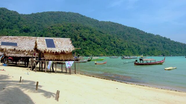 Discover the breathtaking beauty of Surin Island in Thailand as traditional boats glide through crystal-clear waters. Lush greenery surrounds cozy beach huts at the Moken village