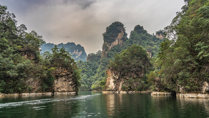 Fototapeta premium Picturesque high mountains surround the calm lake. There is green vegetation on the slopes. Peaks against the sky and clouds. Reflection, ripples on the water. China. Baofeng Lake. Wulingyuan