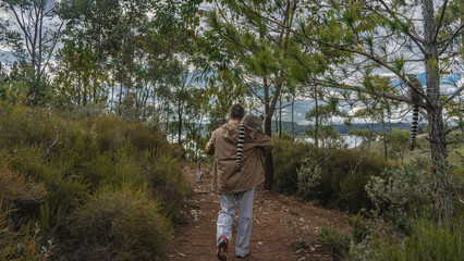 Naklejka premium A man walks along a dirt red soil path. A ring-tailed lemur catta sits on his shoulder. View from the back. Tall grass on the roadsides. Trees against the sky and clouds. Madagascar. Nosy Soa Park