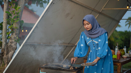 Fototapeta premium Muslim Woman Grilling Meat Outdoors Preparing Food for a Festive Meal