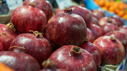 A vibrant close-up of a pomegranate at a bustling market, showcasing its rich red color and textured skin. The blurred background hints at a lively marketplace.
