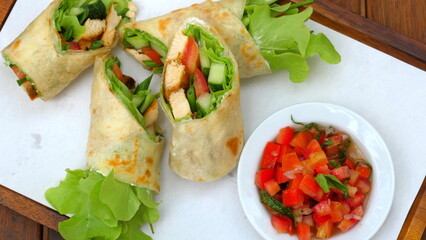 Fresh chicken wraps with lettuce, tomatoes, and cucumbers on white plate next to bowl of diced tomatoes and herbs.