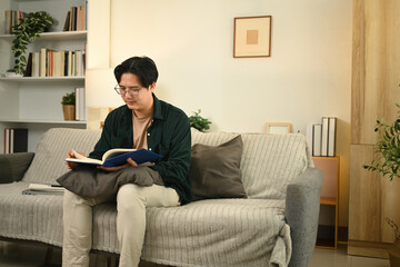 A young man sits on a couch, deeply engrossed in his book. The warm, peaceful setting, complete...