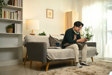 A young Asian man relaxes on a cozy couch, taking a break from work to use his smartphone, he enjoys a moment of digital leisure