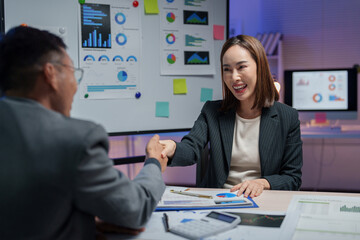 Two business professionals shaking hands in a modern office, discussing charts and graphs. A laptop and documents are visible, emphasizing a collaborative work environment