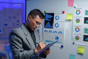 Business analyst in an office setting, reviewing data charts on a whiteboard. The scene is illuminated with blue lighting, emphasizing focus on analytics and strategic planning