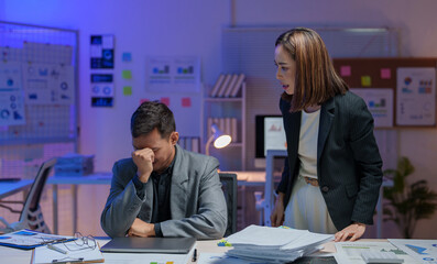 Tense office scene with a frustrated businessman sitting at a desk, while a female colleague stands and speaks emphatically. Papers and charts scattered around suggest a stressful work environment