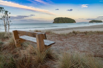 Tranquil Whangamata beach scene. Wooden bench sits on the sand, overlooking the calm ocean and islands. Perfect for relaxation.  Coromandel Peninsula, New Zealand