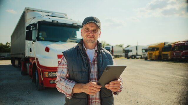 Handsome Caucasian trucker standing in front of long truck. Using tablet device while chatting with customer. Looking at camera and smiling with joy. Wearing baseball cap and warm vest.