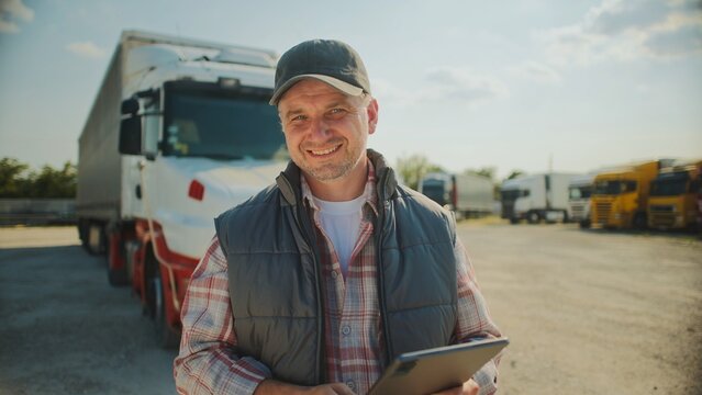 Handsome Caucasian trucker standing in front of long truck. Using tablet device while chatting with customer. Looking at camera and smiling with joy. Wearing baseball cap and warm vest.