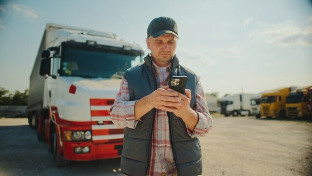 Focused Caucasian male standing in front of his truck while using smartphone. Looking at screen. Pressing with fingers on touch screen. Confirming finished delivery. Sunny weather outside.