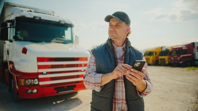 Focused Caucasian male standing in front of his truck while using smartphone. Looking at screen. Pressing with fingers on touch screen. Confirming finished delivery. Sunny weather outside.