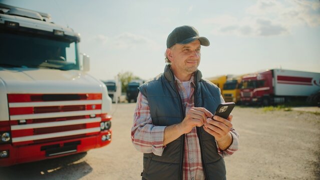 Focused Caucasian male standing in front of his truck while using smartphone. Looking at screen. Pressing with fingers on touch screen. Confirming finished delivery. Sunny weather outside.