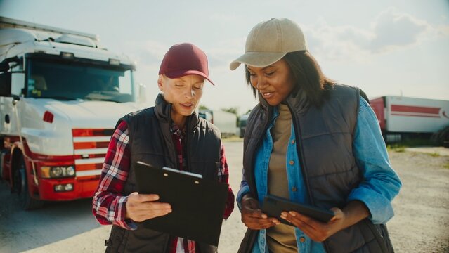 Two women standing in middle of parking lot. Talking with each other. Looking at tablet device with information on display. Modern worker utilizing technology to stay connected and organized.