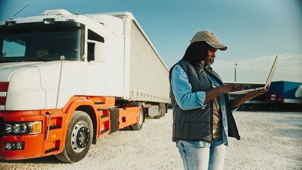 Side camera view of African American woman using laptop while standing next to long truck. Typing...
