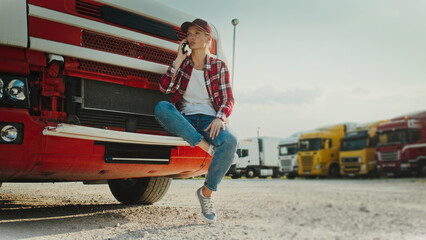 Charming Caucasian woman sitting on drop bench of her truck. Wearing checkered shirt and jeans. Talking on smartphone. Communicating remotely with friend of relative. Trucker resting in middle of trip