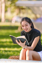 Obraz premium Young woman sitting on a hay bale in a park, smiling and immersed in a book, relishing the tranquil atmosphere while enjoying the beauty of nature around her